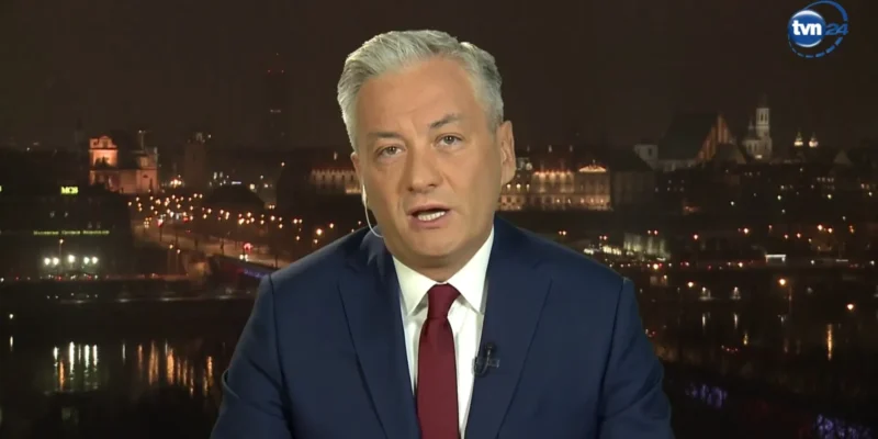 Dziennikarz w granatowym garniturze i czerwonym krawacie przemawia do kamery na tle nocnego miasta; w prawym górnym rogu widoczny jest logotyp TVN24.