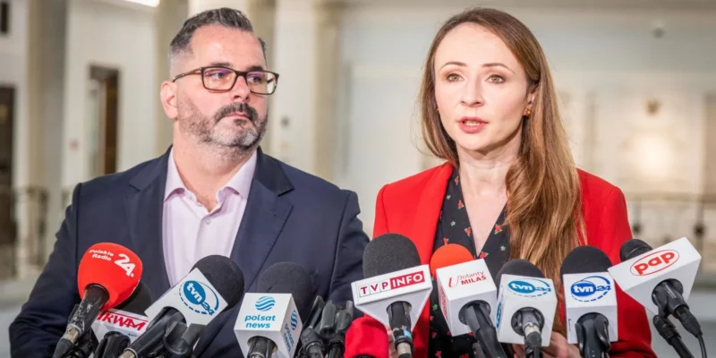 Na zdjęciu widoczni są dwoje mówiących do mediów polityków, otoczonych mikrofonami z logo różnych stacji telewizyjnych i radiowych, w serdecznej atmosferze konferencji prasowej.