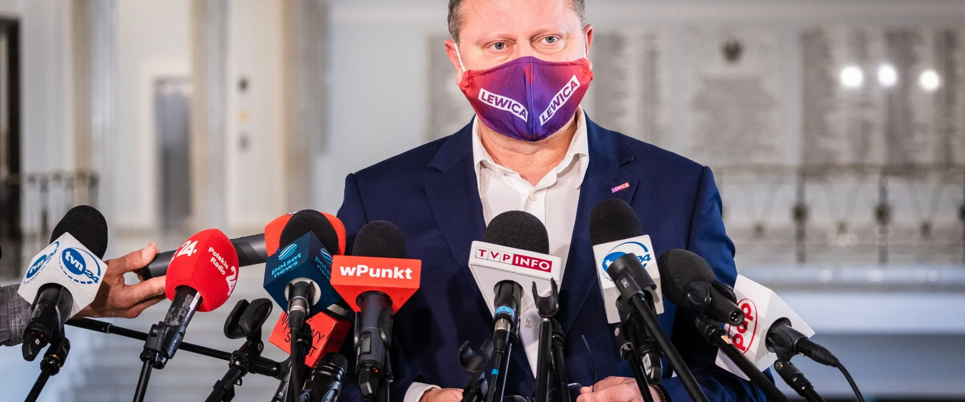 Polityk w granatowym garniturze i fioletowo-czerwonej masce z napisem LEWICA przemawia do mediów podczas konferencji prasowej, stojąc przed licznymi mikrofonami.