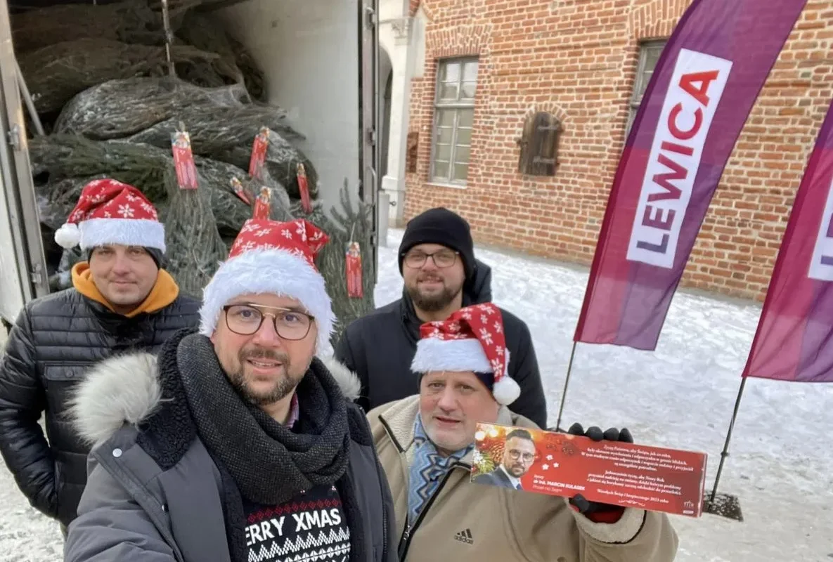 Na zdjęciu czterech mężczyzn w czapkach świątecznych pozuje przed ciężarówką z choinkami, trzymając ulotkę w zimowej scenerii.