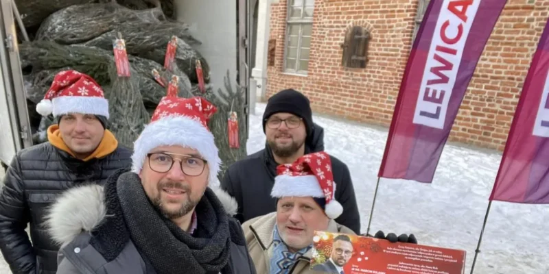 Na zdjęciu czterech mężczyzn w czapkach świątecznych pozuje przed ciężarówką z choinkami, trzymając ulotkę w zimowej scenerii.