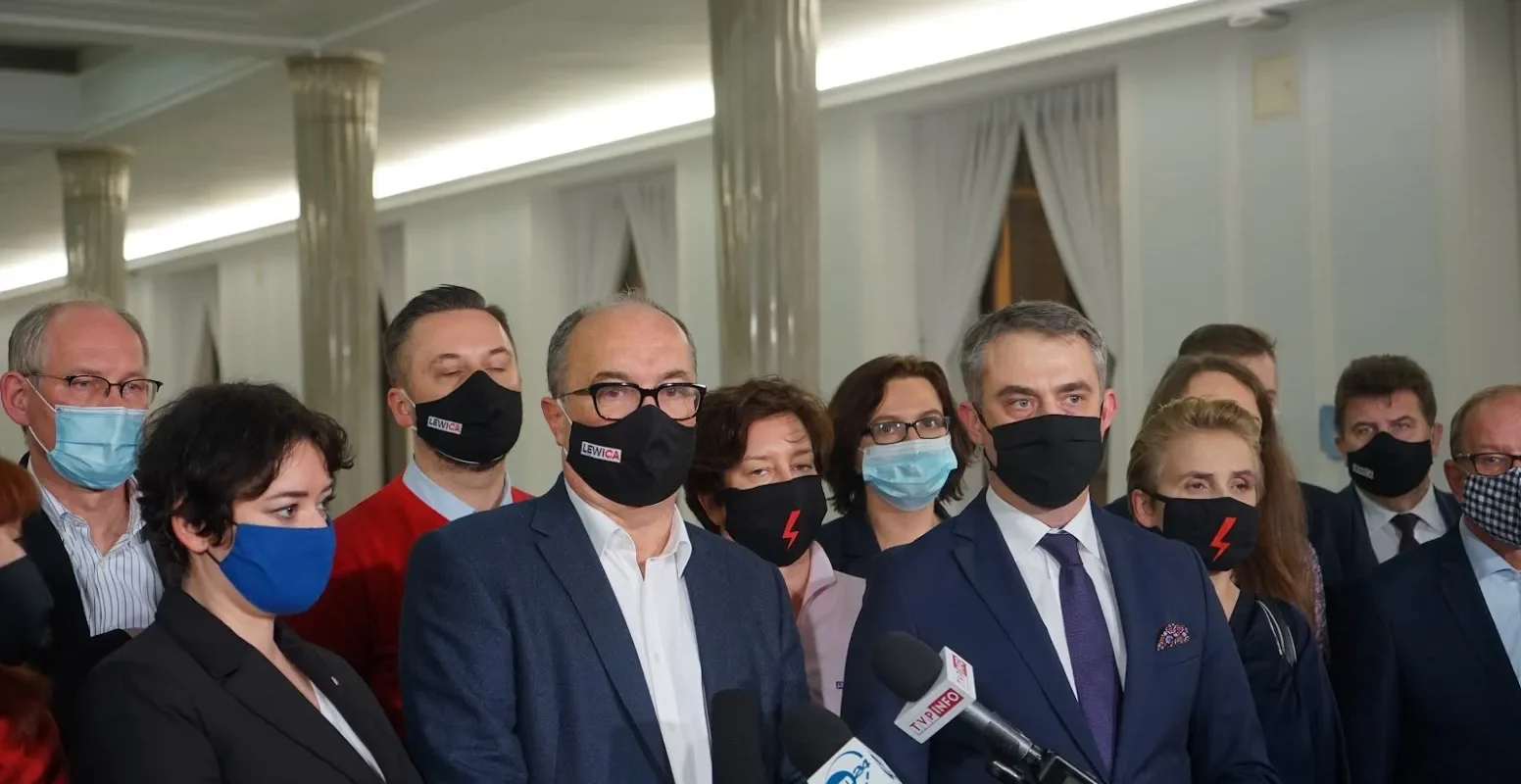 Grupa polityków w garniturach i maseczkach stoi przy mikrofonach podczas konferencji prasowej w jasnej, eleganckiej sali.