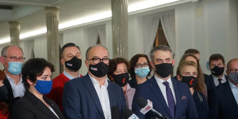 Grupa polityków w garniturach i maseczkach stoi przy mikrofonach podczas konferencji prasowej w jasnej, eleganckiej sali.