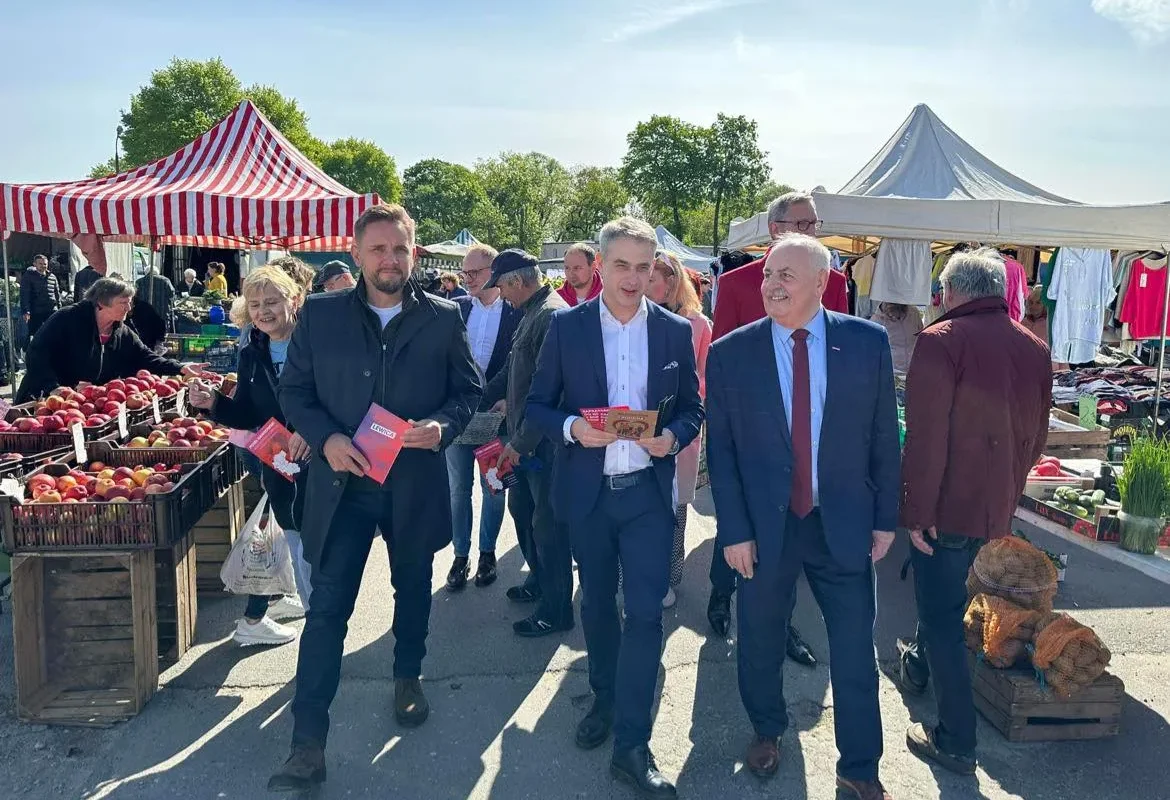 Grupa osób spaceruje po targowisku w Kujawsko-Pomorskiem, pod hasłem „Bezpieczna Rodzina”, trzymając ulotki, a w tle stoją stoiska z owocami i warzywami.