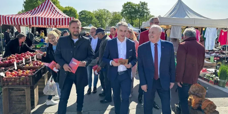 Grupa osób spaceruje po targowisku w Kujawsko-Pomorskiem, pod hasłem „Bezpieczna Rodzina”, trzymając ulotki, a w tle stoją stoiska z owocami i warzywami.