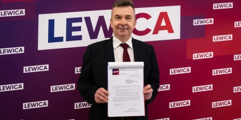 Mężczyzna w garniturze trzyma przed sobą dokument na tle ściany z logotypami Lewicy.