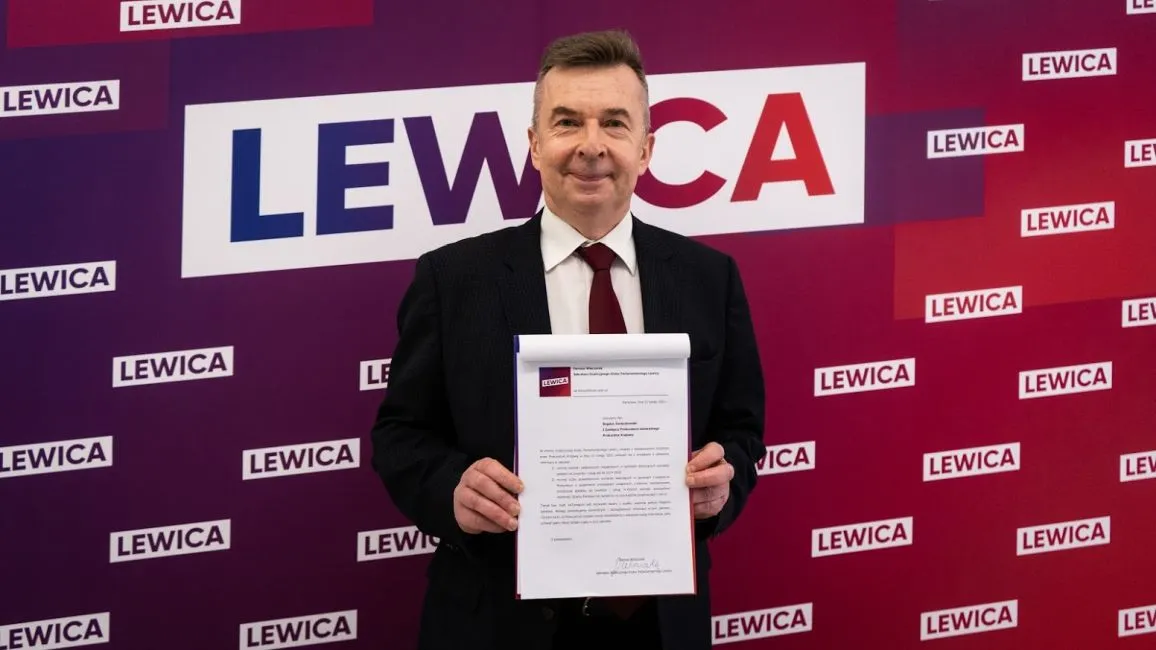 Mężczyzna w garniturze trzyma przed sobą dokument na tle ściany z logotypami Lewicy.