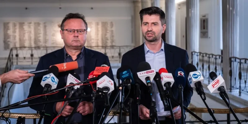 Na zdjęciu dwóch mężczyzn stoi obok siebie przed mikrofonami, jeden z nich mówi, podczas gdy drugi słucha, w tle widoczna jest elegancka architektura.