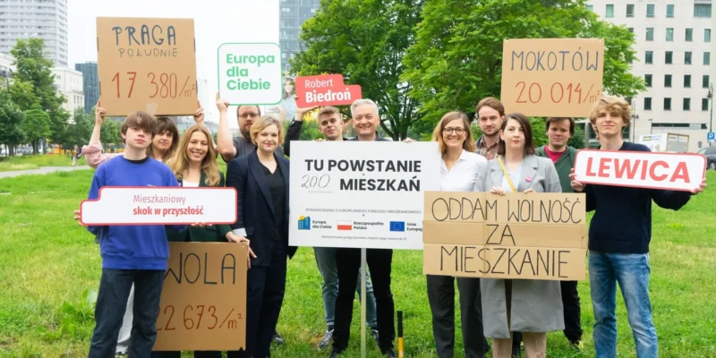 Grupa demonstrantów w parku trzyma kartonowe plakaty o mieszkaniach i kredycie 0 procent, protestując przeciw dopłatom dla banków i deweloperów.
