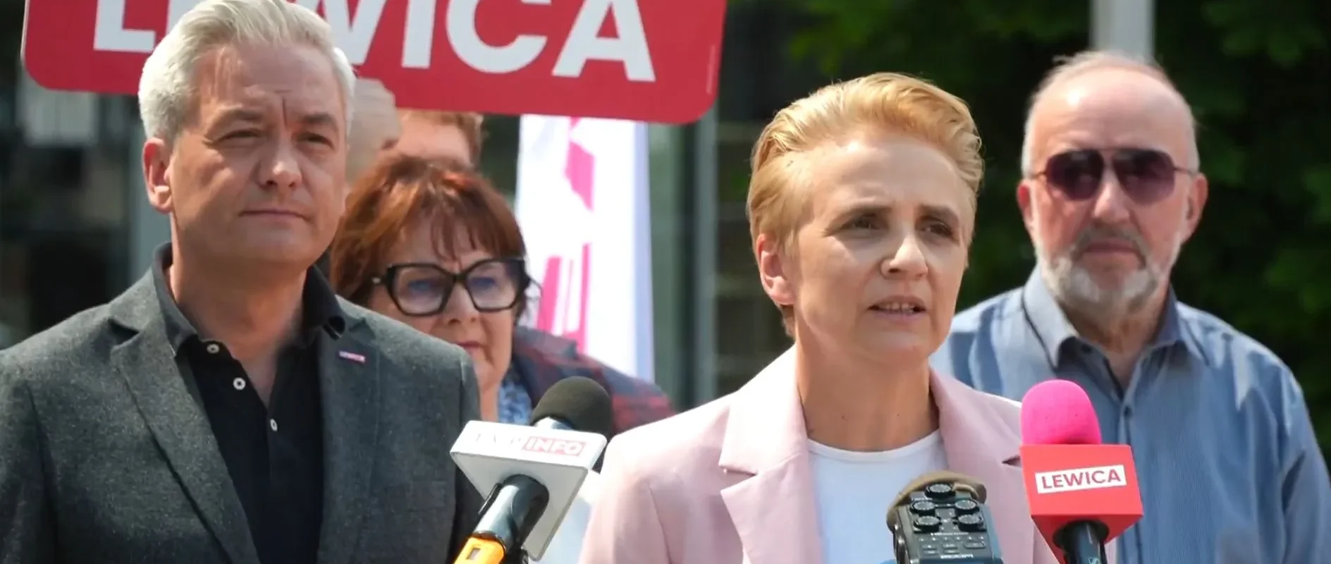 Konferencja prasowa Lewicy: blond kobieta w różowym żakiecie przemawia do mikrofonów, stoją obok niej mężczyzna w ciemnym garniturze, a w tle widać czerwony znak Lewicy.