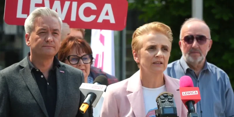 Konferencja prasowa Lewicy: blond kobieta w różowym żakiecie przemawia do mikrofonów, stoją obok niej mężczyzna w ciemnym garniturze, a w tle widać czerwony znak Lewicy.