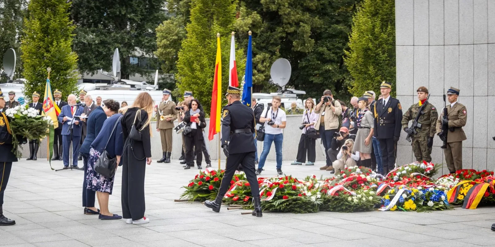 Uroczystość upamiętniająca 79. rocznicę Powstania Warszawskiego, podczas której żołnierze w mundurach składają wieńce, wokół stoją flagi, a fotografowie dokumentują wydarzenie.