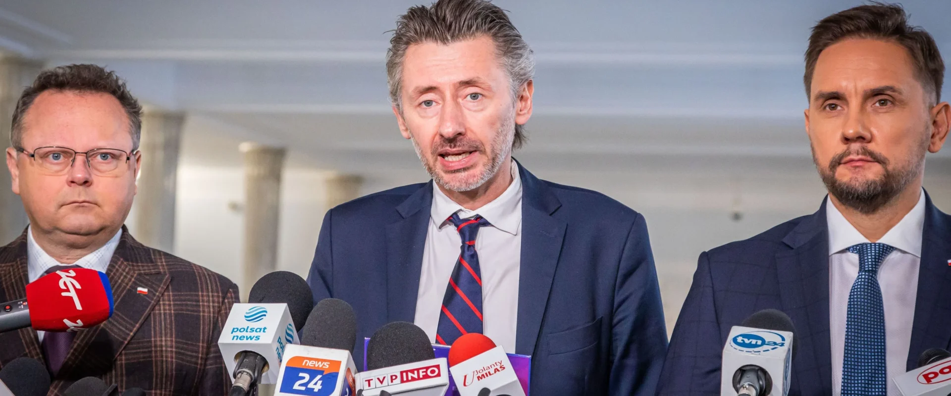 Na zdjęciu trzech mężczyzn stoi przy mikrofonach podczas konferencji prasowej.