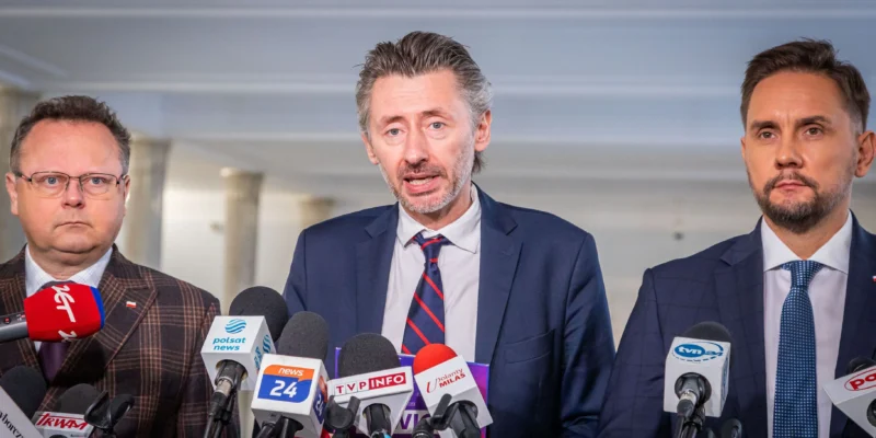 Na zdjęciu trzech mężczyzn stoi przy mikrofonach podczas konferencji prasowej.