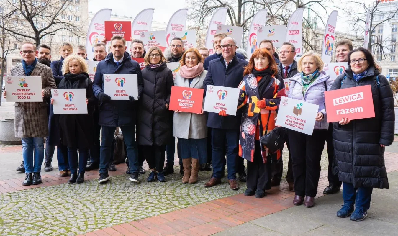 Grupa osób trzymająca transparenty z hasłami o wsparciu socjalnym i publicznych usługach, pozująca do zdjęcia na ulicy.