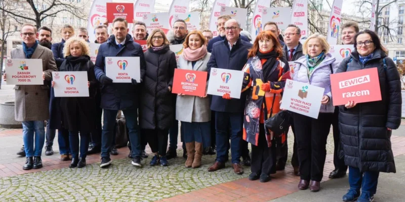 Grupa osób trzymająca transparenty z hasłami o wsparciu socjalnym i publicznych usługach, pozująca do zdjęcia na ulicy.