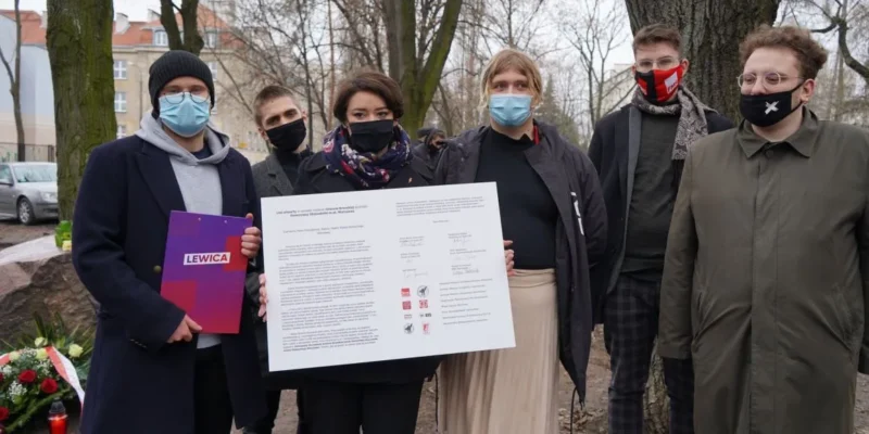 Grupa osób w maseczkach stoi na zewnątrz i prezentuje duży plakat z tekstem i podpisami, obok kwiatów złożonych przy pomniku podczas rocznicy śmierci Jolanty Brzeskiej.