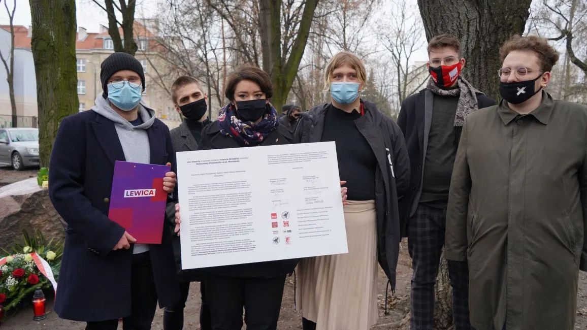 Grupa osób w maseczkach stoi na zewnątrz i prezentuje duży plakat z tekstem i podpisami, obok kwiatów złożonych przy pomniku podczas rocznicy śmierci Jolanty Brzeskiej.
