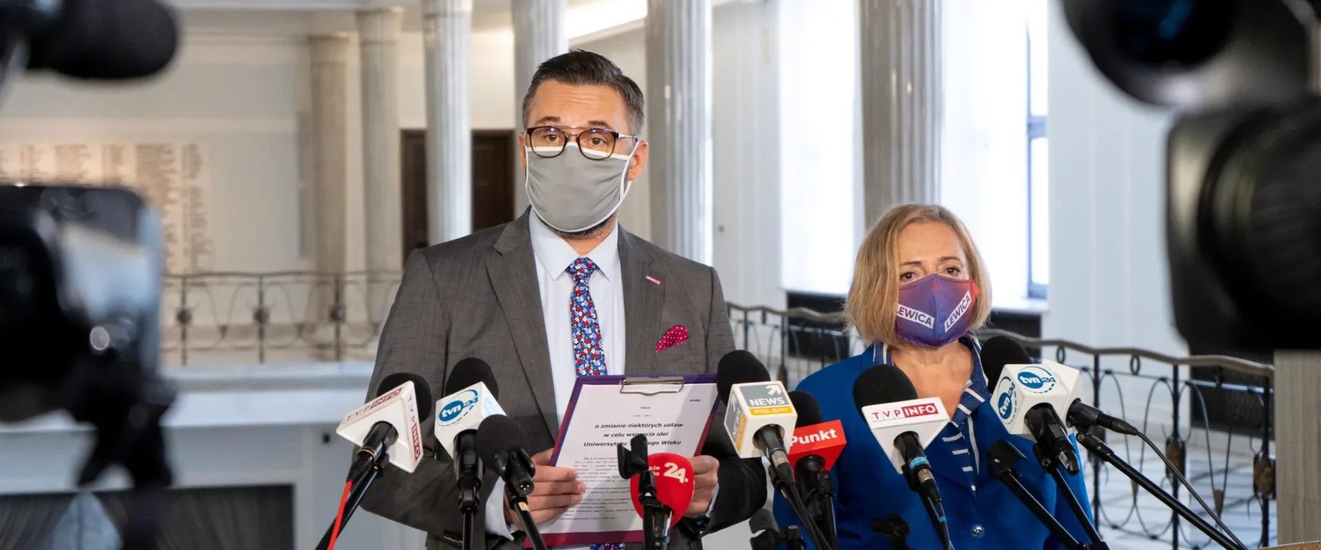 Dwóch przedstawicieli Lewicy na konferencji prasowej w holu z kolumnami, mężczyzna w garniturze z maseczką prezentuje dokument dotyczący ustawy o Uniwersytecie Trzeciego Wieku w każdym powiecie, obok niego kobieta w niebieskim stroju z maseczką z napisem Lewica, otoczeni licznymi mikrofonami.