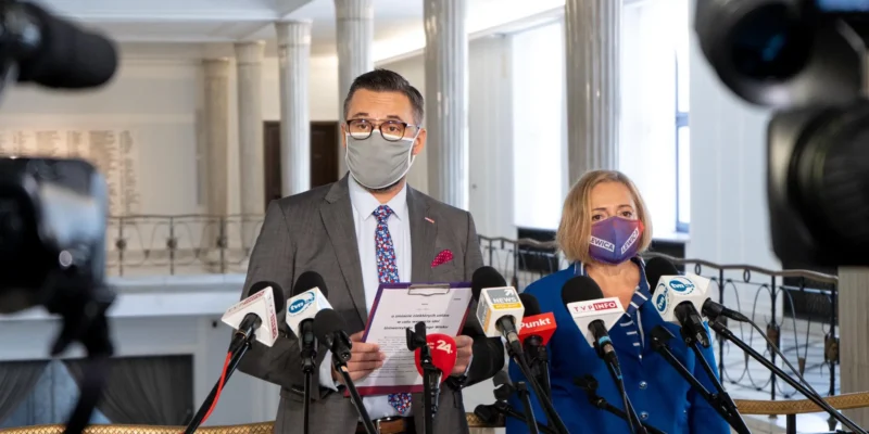 Dwóch przedstawicieli Lewicy na konferencji prasowej w holu z kolumnami, mężczyzna w garniturze z maseczką prezentuje dokument dotyczący ustawy o Uniwersytecie Trzeciego Wieku w każdym powiecie, obok niego kobieta w niebieskim stroju z maseczką z napisem Lewica, otoczeni licznymi mikrofonami.