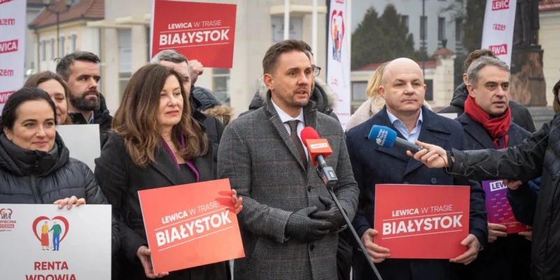 Na zdjęciu grupa osób z partii Lewica prowadzi konferencję prasową w Białymstoku, trzymając transparenty i plakaty dotyczące aktu politycznego.