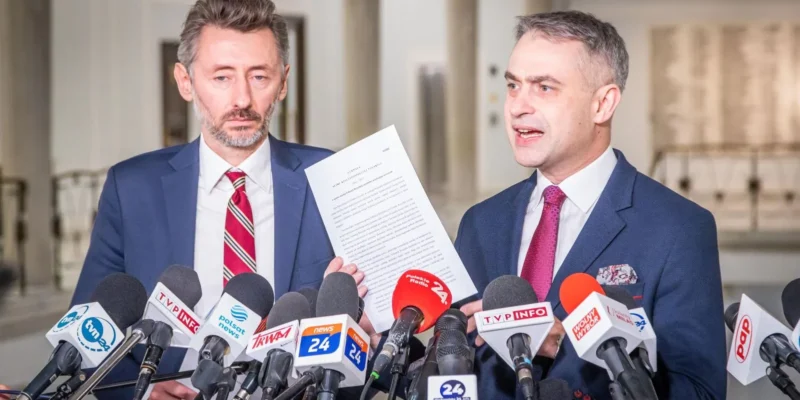 Na zdjęciu dwaj mężczyźni występują na konferencji prasowej, trzymając dokument, w otoczeniu mikrofonów medialnych.