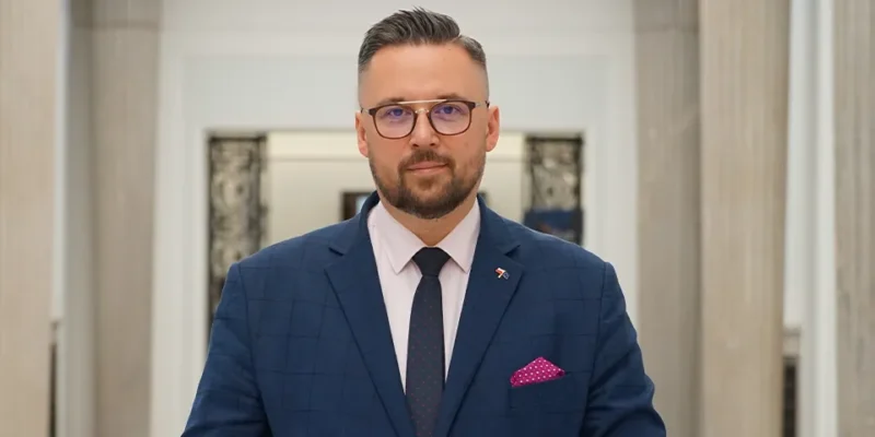 Mężczyzna w eleganckim niebieskim garniturze stoi w przestronnej hali z kolumnami.