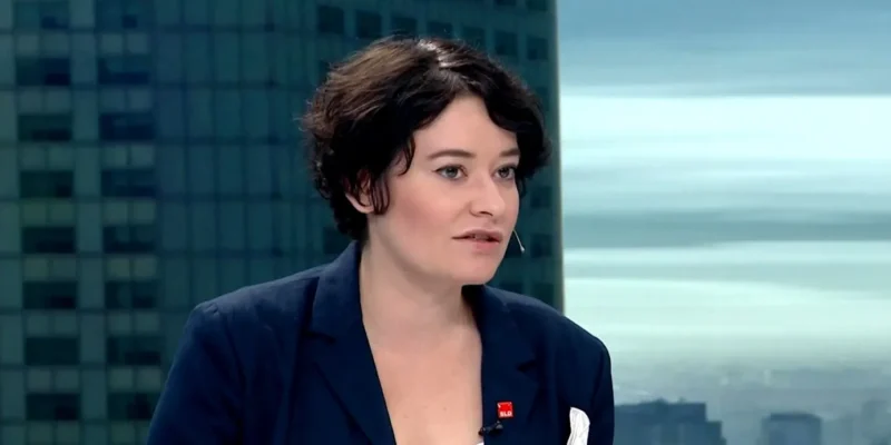 Kobieta o krótkich ciemnych włosach w granatowym żakiecie rozmawia w studiu telewizyjnym, z mikrofonem krawatowym, na tle miejskiego krajobrazu.