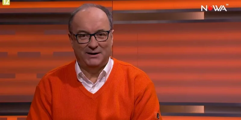 Mężczyzna w okularach ubrany w pomarańczowy sweter siedzi w studiu telewizyjnym, w prawym górnym rogu widoczne logo Nowa TV.