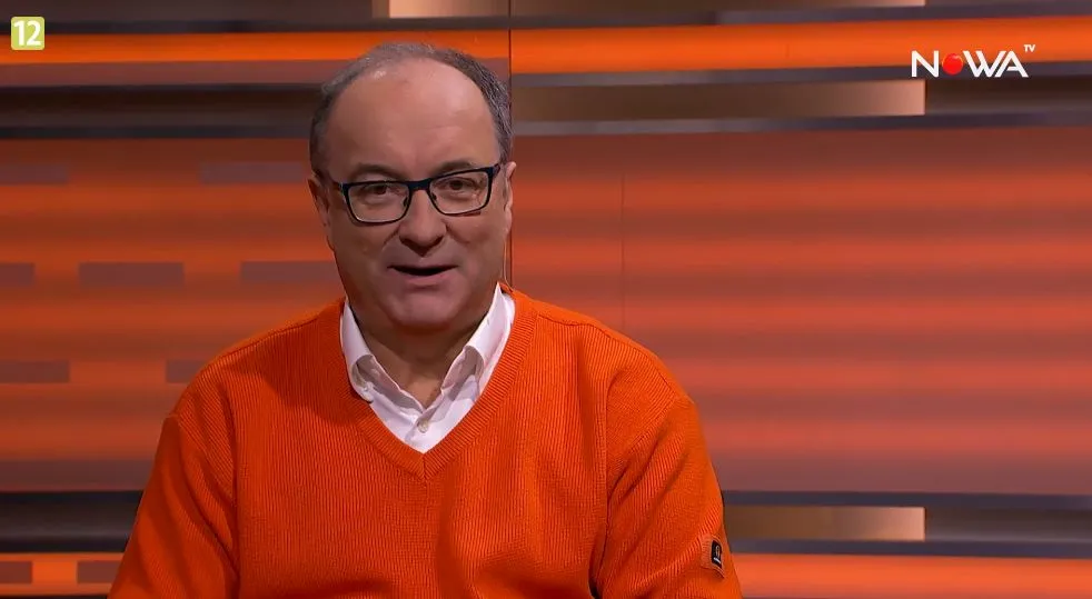 Mężczyzna w okularach ubrany w pomarańczowy sweter siedzi w studiu telewizyjnym, w prawym górnym rogu widoczne logo Nowa TV.