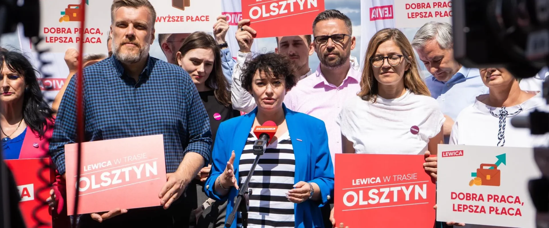 Grupa polityków Lewicy na wiecu w Olsztynie trzyma plakaty z hasłami „Lewica w trasie Olsztyn” i „Dobra praca, lepsza płaca”, a kobieta w niebieskim żakiecie przemawia do mikrofonu.