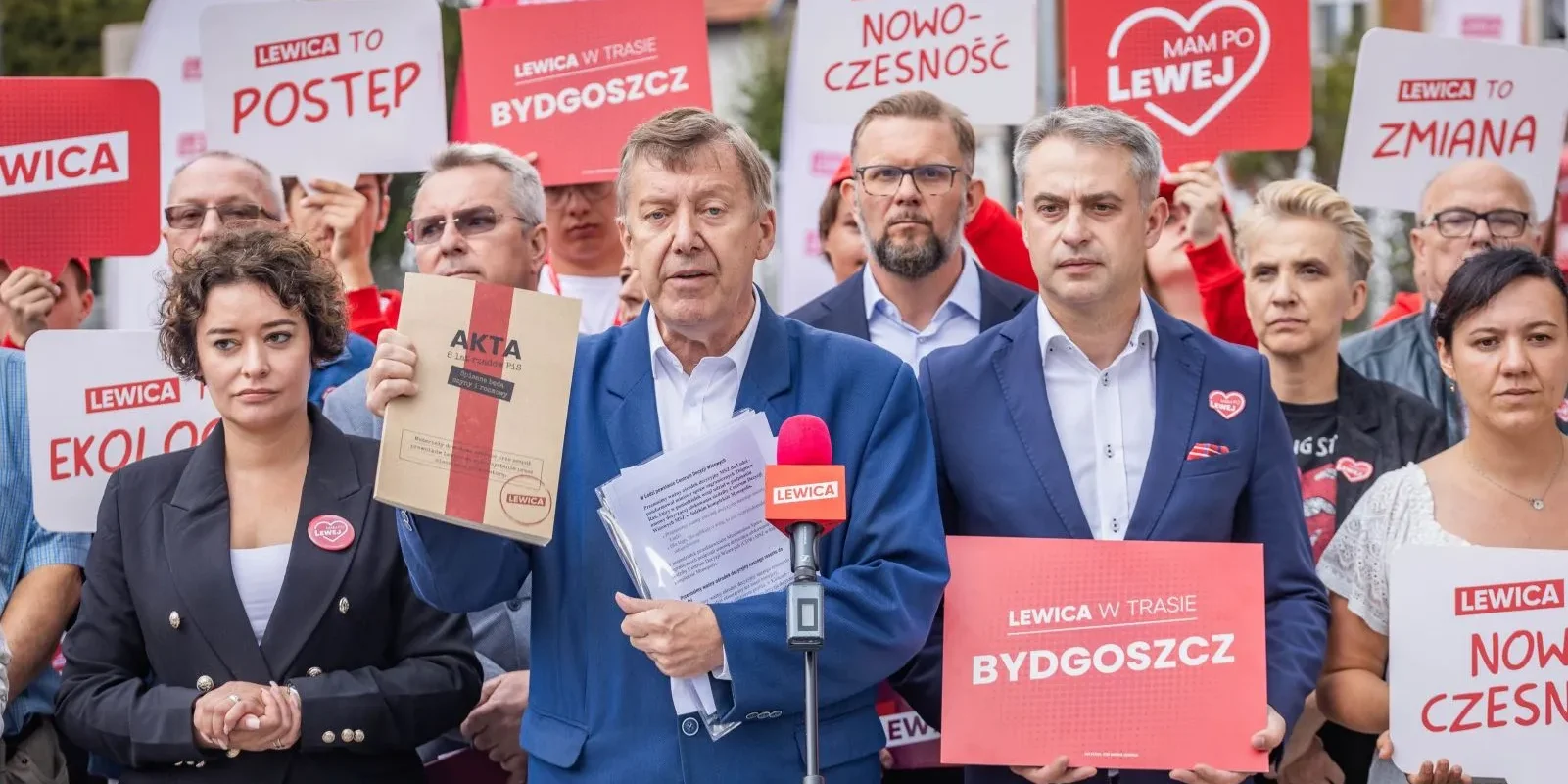 Grupa zwolenników Lewicy na wiecu z plakatami i hasłami Lewica; na pierwszym planie mężczyzna w niebieskim garniturze przemawia do mikrofonu, trzymając karton z napisem AKTA i dokumenty.