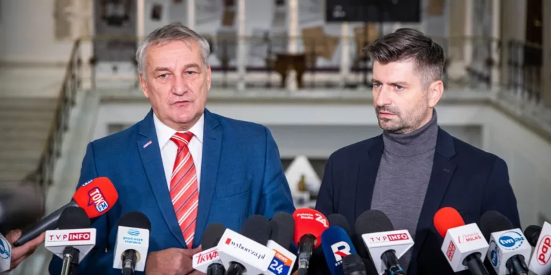 Na zdjęciu widać dwóch mężczyzn stojących przed mikrofonami i kamerami w obszernym wnętrzu, prawdopodobnie podczas konferencji prasowej.