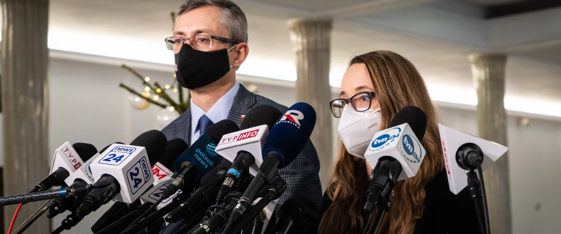 Na zdjęciu widać dwóch mówców przygotowujących się do wystąpienia przed zgromadzeniem mediów, otoczonych przez mikrofony i kamery.