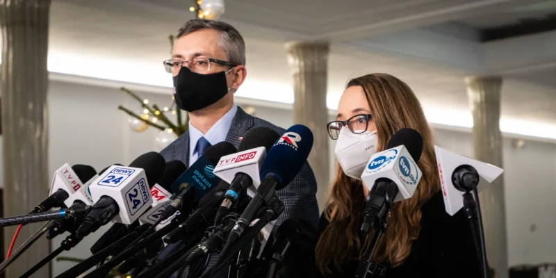 Na zdjęciu widać dwóch mówców przygotowujących się do wystąpienia przed zgromadzeniem mediów, otoczonych przez mikrofony i kamery.