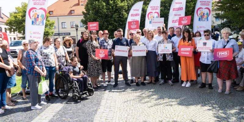 Grupa ludzi różnych wieku stoi na placu podczas wydarzenia, trzyma plakaty i flagi Lewica i Razem, a w tle widać banery z hasłem „Bezpieczna Rodzina”.