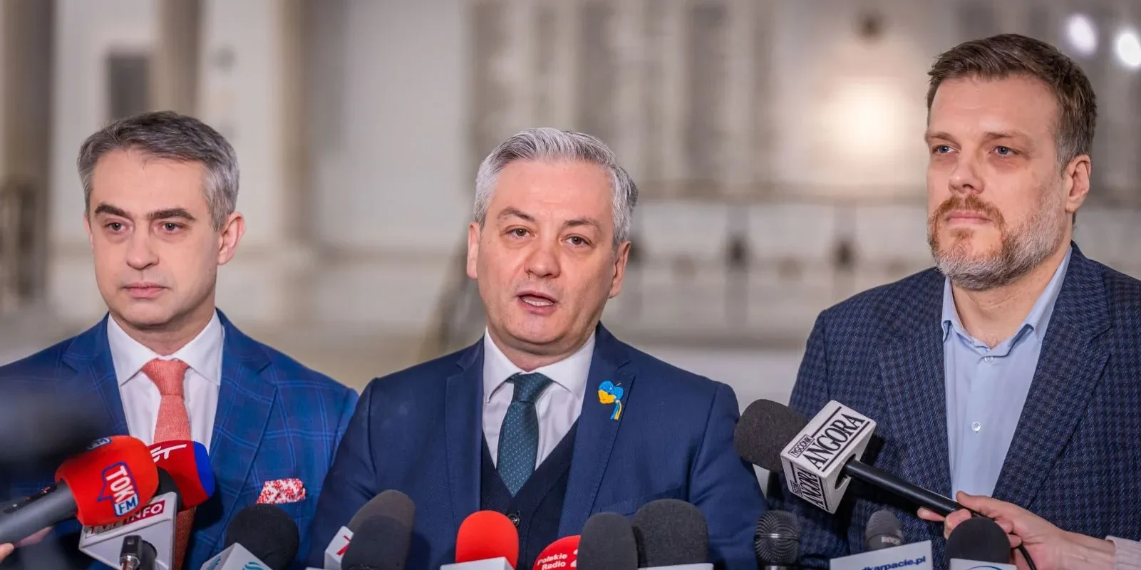 Na zdjęciu widoczny jest trzech mężczyzn w garniturach, którzy stoją przed mikrofonami podczas konferencji prasowej w jasnym wnętrzu.