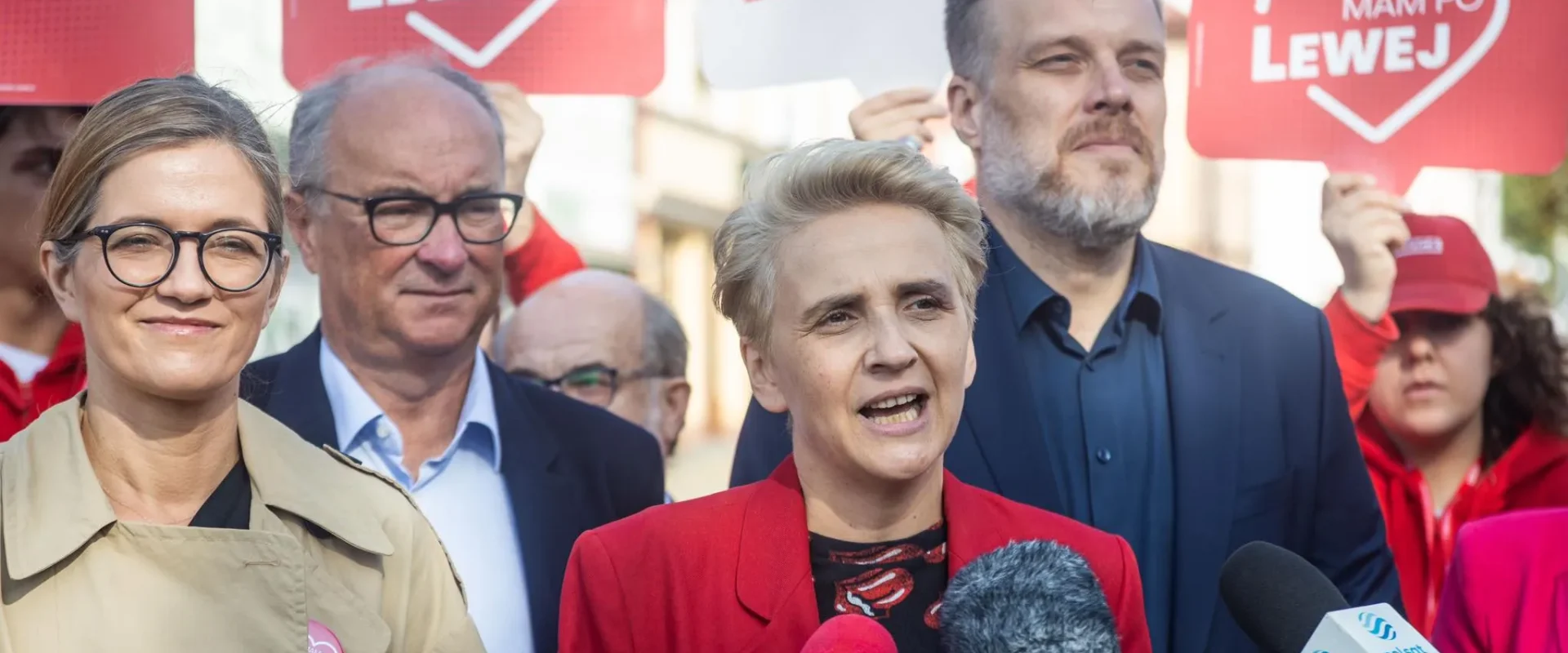 Kobieta w czerwonej marynarce przemawia do mediów podczas wiecu Lewicy, w tle ludzie trzymają plakaty z napisem LEWICA.