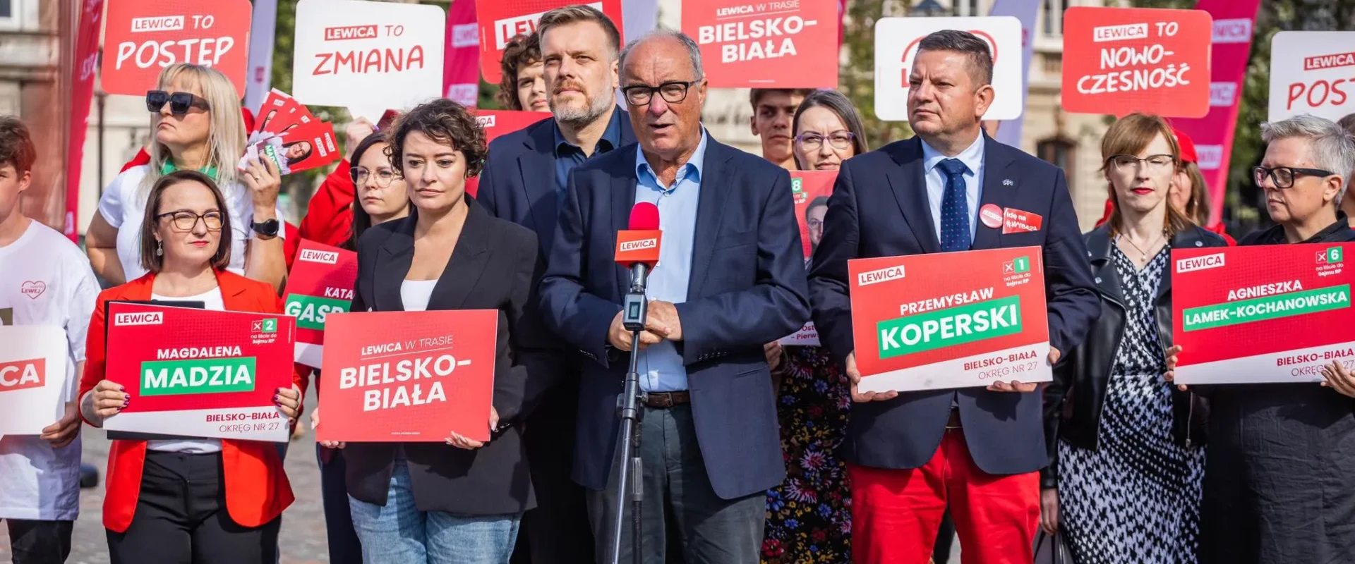 Grupa działaczy Lewicy na wiecu wyborczym trzyma czerwono-białe plakaty z nazwiskami kandydatów, podczas gdy na środku mężczyzna przemawia do mikrofonu.