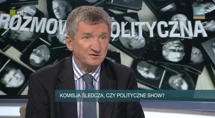 Mężczyzna w garniturze w studiu telewizyjnym z paskiem „KOMISJA ŚLEDCZA, CZY POLITYCZNE SHOW?” i tłem „ROZMOWA POLITYCZNA”.