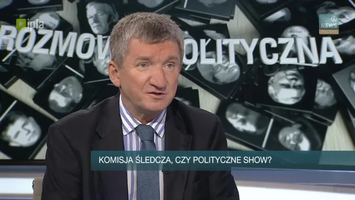 Mężczyzna w garniturze w studiu telewizyjnym z paskiem „KOMISJA ŚLEDCZA, CZY POLITYCZNE SHOW?” i tłem „ROZMOWA POLITYCZNA”.