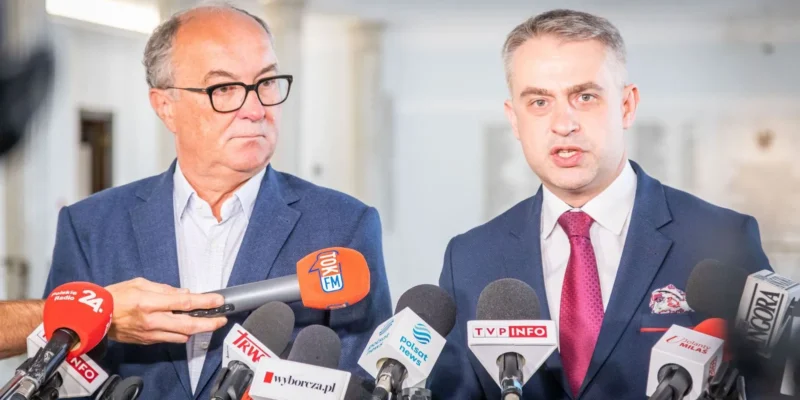 Dwóch mężczyzn w garniturach udziela konferencji prasowej, stojąc wśród licznych mikrofonów.