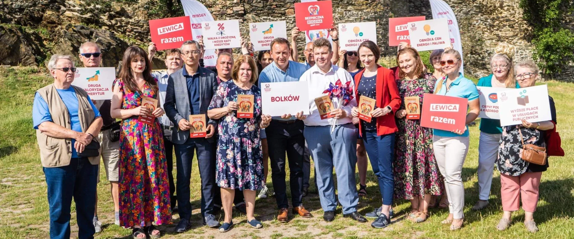 Grupa posłów Lewicy wraz z mieszkańcami stoi na zewnątrz przed kamienną ścianą, trzymając plakaty i ulotki związane z trasą Bezpieczna rodzina podczas wizyty w Lubuskiem i na Dolnym Śląsku.