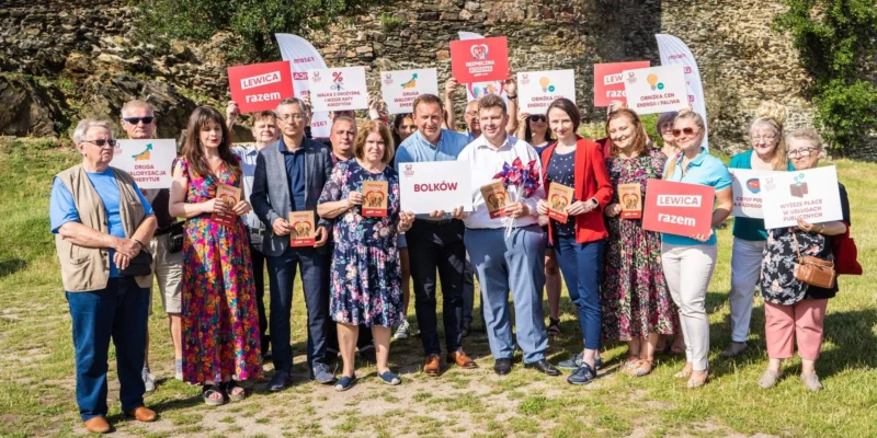 Grupa posłów Lewicy wraz z mieszkańcami stoi na zewnątrz przed kamienną ścianą, trzymając plakaty i ulotki związane z trasą Bezpieczna rodzina podczas wizyty w Lubuskiem i na Dolnym Śląsku.