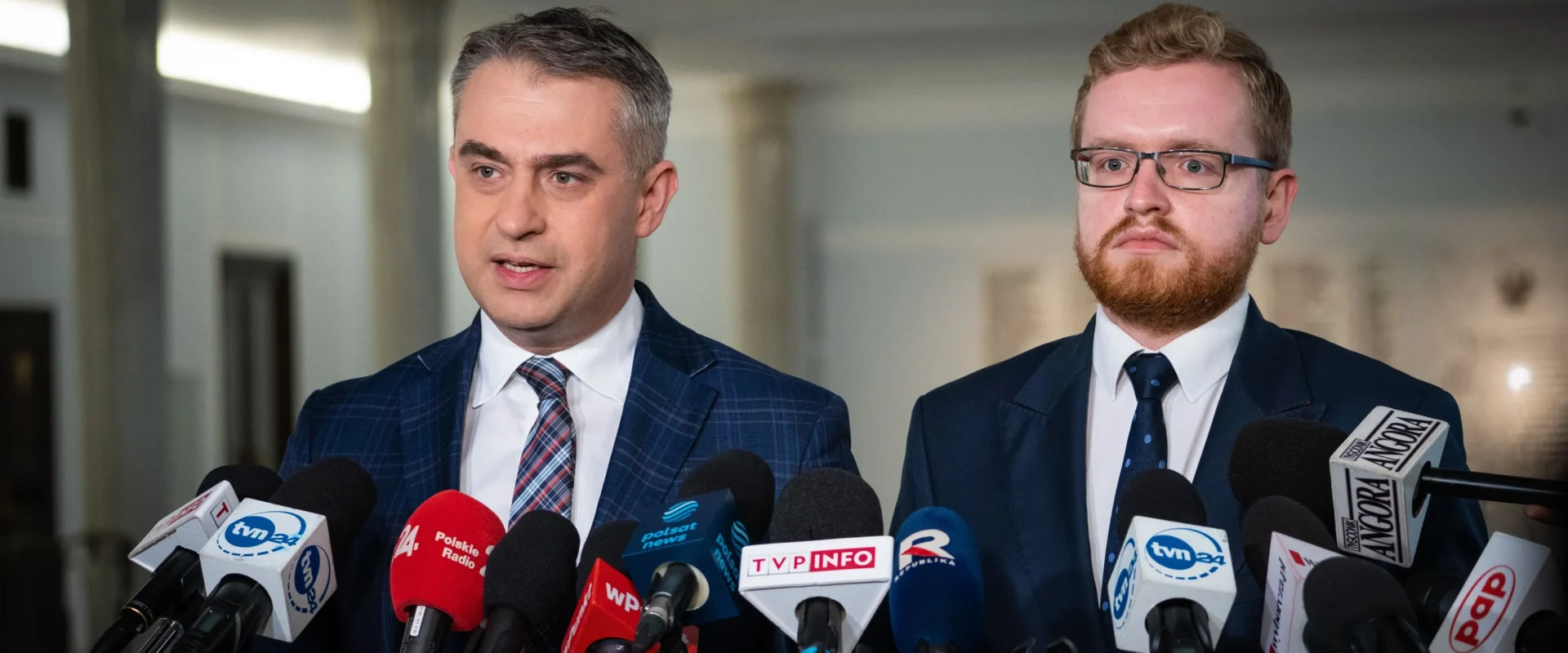 Dwóch mężczyzn w garniturach przemawia na konferencji prasowej, stojąc przed licznymi mikrofonami mediów.