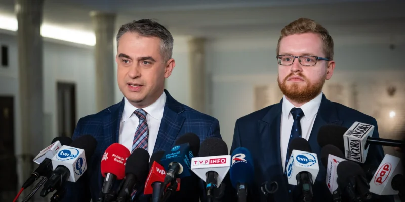 Dwóch mężczyzn w garniturach przemawia na konferencji prasowej, stojąc przed licznymi mikrofonami mediów.