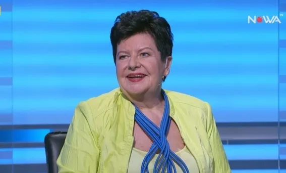 Uśmiechnięta kobieta w jasnej żółtej marynarce i niebieskim ozdobnym sznurze przy szyi siedzi w studiu telewizyjnym na tle niebieskich pasów.