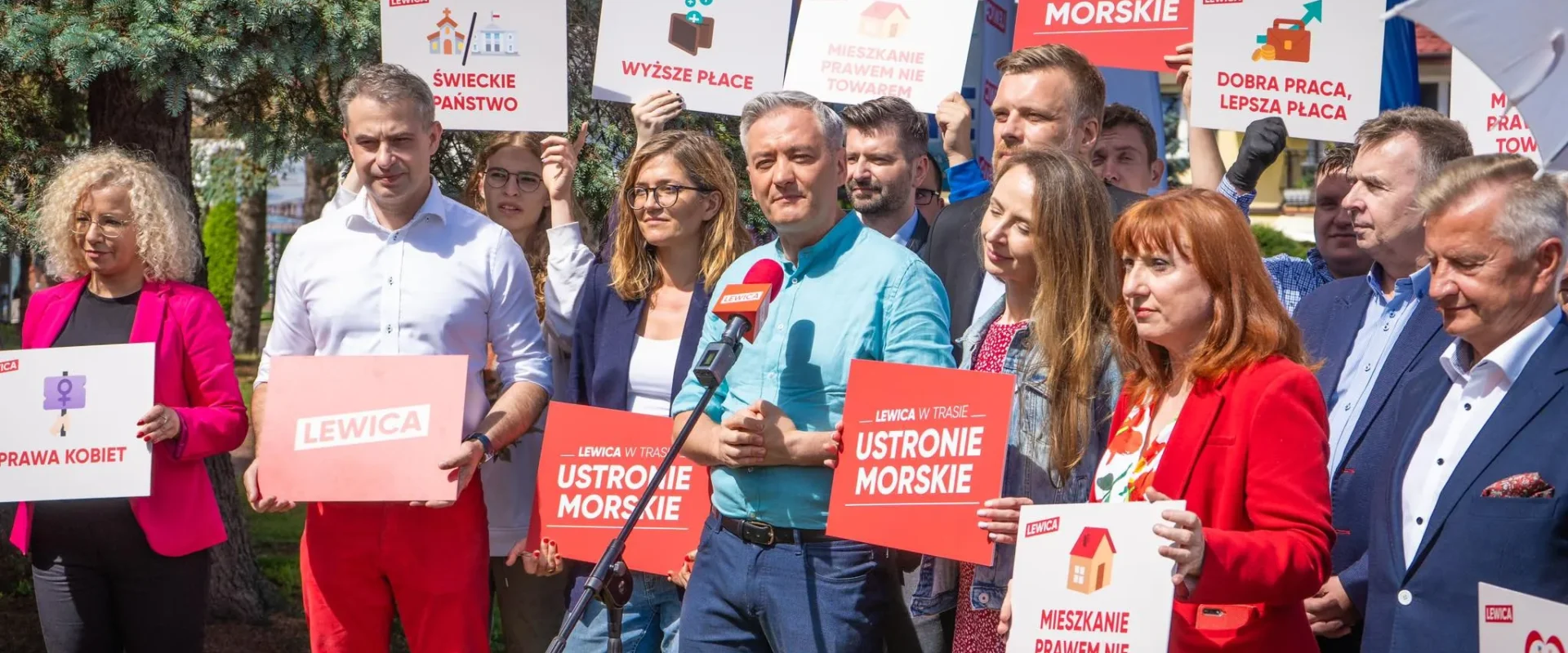 Grupa polityków Lewicy podczas konferencji prasowej trzyma plakaty z hasłami, a mężczyzna w turkusowej koszuli przemawia do mikrofonu.