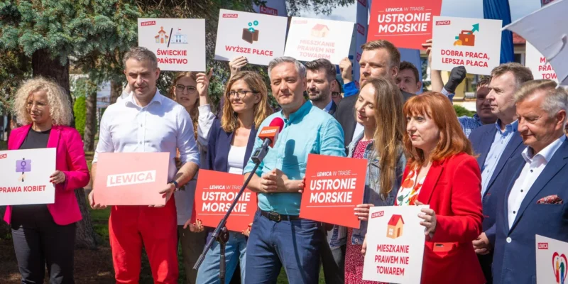 Grupa polityków Lewicy podczas konferencji prasowej trzyma plakaty z hasłami, a mężczyzna w turkusowej koszuli przemawia do mikrofonu.