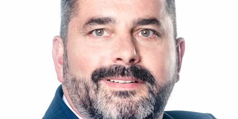 Mężczyzna w garniturze z jasno niebieską koszulą i czerwoną krawatem, uśmiechający się na tle białym.